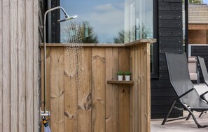Una ducha exterior de madera con un grifo de metal, un estante pequeño con dos plantas en macetas y una silla negra en un patio.