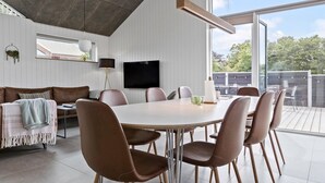 Ein moderner Essbereich mit einem runden Tisch und sechs Stühlen, einem flachbildfernseher an der Wand und Blick ins Freie durch große Fenster.