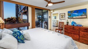 A hotel room with a large bed, a ceiling fan, a wooden dresser, and a view of the outdoors through large windows. - Spectacular Ocean Front, Garden Level, Corner Unit, Stunning Views for 4 in Kona (Kailua Kona)