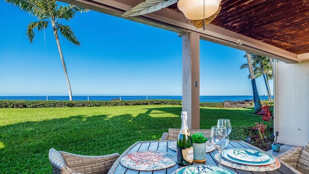 An outdoor dining area with a view of the ocean, a table set with plates and glasses, and a bottle of champagne. - Spectacular Ocean Front, Garden Level, Corner Unit, Stunning Views for 4 in Kona (Kailua Kona)