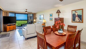A modern living room with a large flat-screen TV, a dining table with chairs, and a view of the ocean through large windows. - Spectacular Ocean Front, Garden Level, Corner Unit, Stunning Views for 4 in Kona (Kailua Kona)