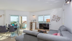 Una sala de estar moderna con un sofá gris, un comedor con sillas y una mesa, un televisor y un balcón con vistas al paisaje.