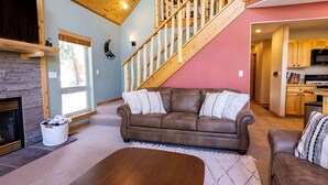 A living room with a stone fireplace, a brown leather sofa, a wooden coffee table, and a staircase leading to an upper level. - Snow Banks Lodge, Beautiful Home in Angel Fire (Angel Fire)