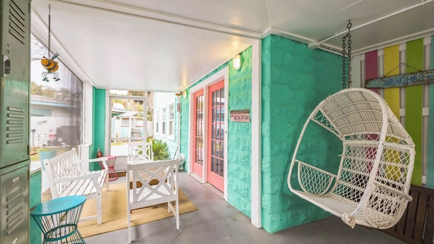 Een overdekt terras met witte stoelen en een hangstoel, een blauwgroene muur, en een bordje met de tekst 'BEACH HOUSE'.