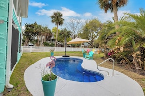 A swimming pool with a curved edge, surrounded by a white deck. There are chairs and a table in the background, and a palm tree is visible. - After Dune Delight by Tybee Vacation Rentals (Tybee Island)