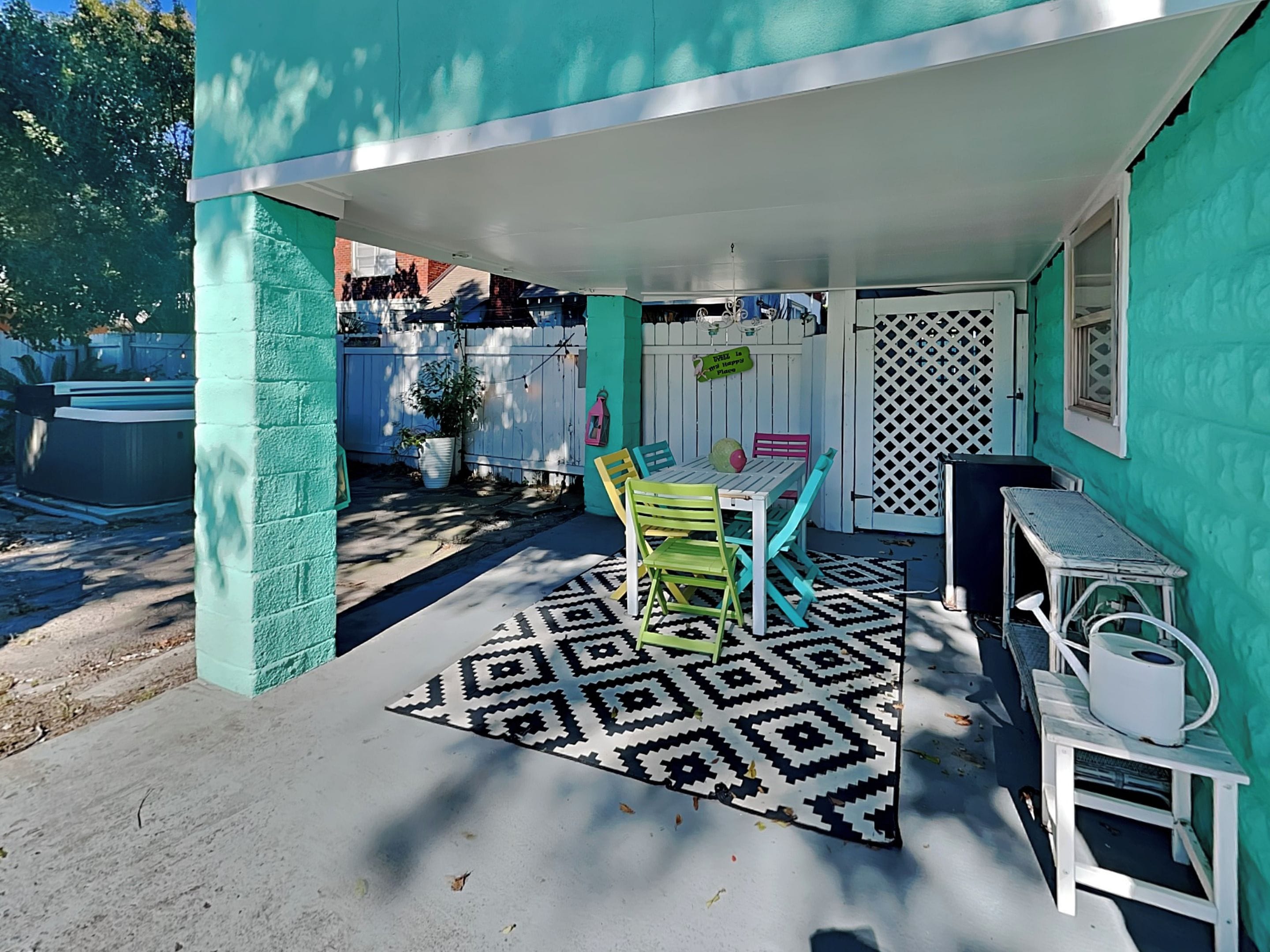 Ein Terrassenbereich im Freien mit Tisch und Stühlen, einem gemusterten Teppich und einem weißen Zaun im Hintergrund.