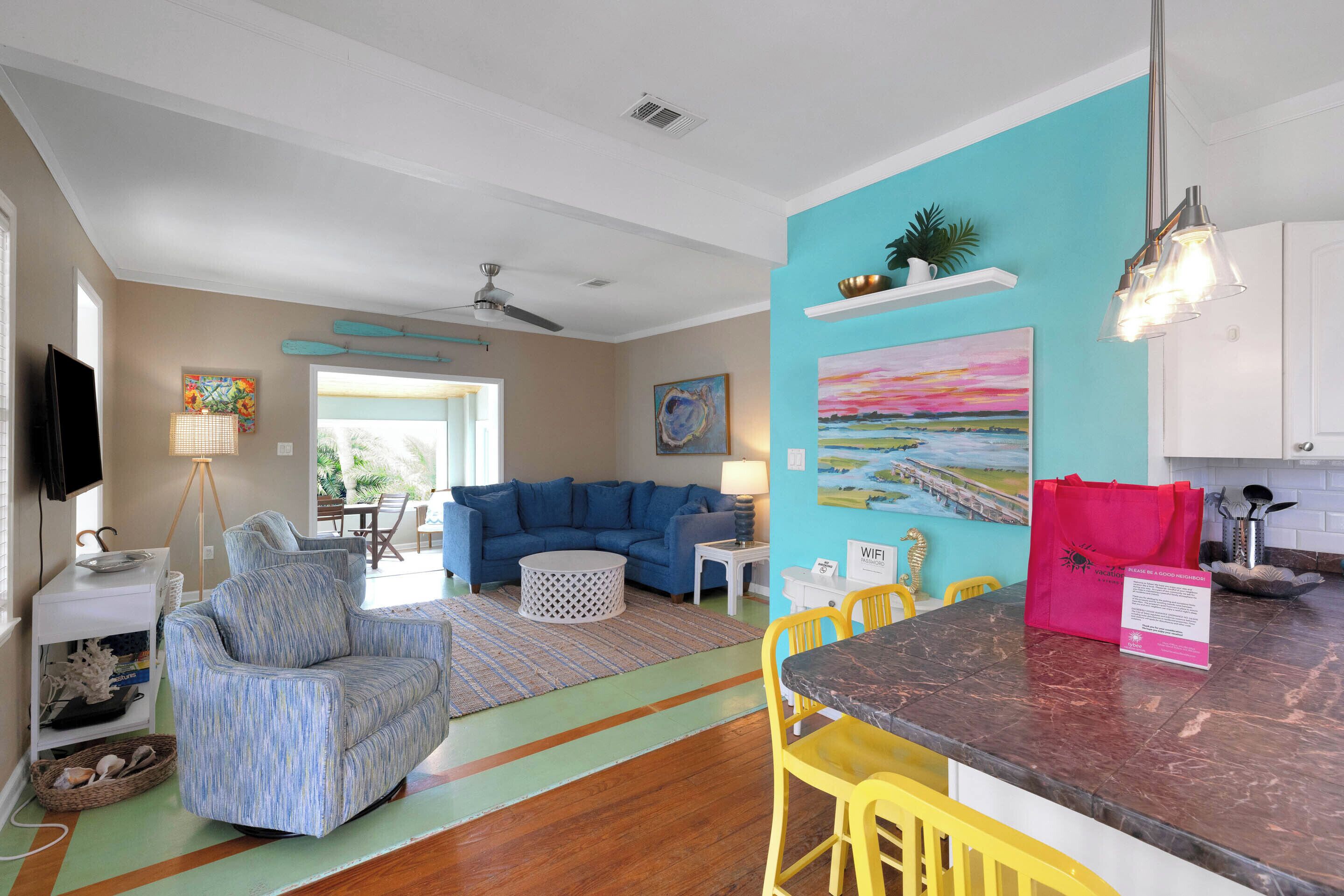 Una sala de estar moderna con un sofá azul, una mesita redonda pequeña y una silla amarilla. Se observa una cocina con encimera de mármol y un bolso rojo encima.