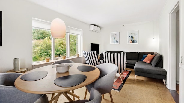 A modern living room with a round wooden table, grey upholstered chairs, a grey sofa with colorful cushions, and a large window offering a view of greenery. - Cozy home in Nørre Nebel with WiFi (Nørre Nebel)