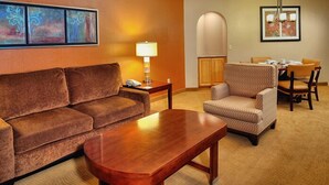 Quarto de hotel com um sofá marrom, uma mesa de centro de madeira, uma poltrona estampada, uma mesa de jantar com cadeiras e um abajur.