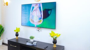 Un téléviseur à écran plat fixé à un mur blanc, une horloge au-dessus, et deux vases avec des fleurs jaunes placés sur une console sombre.
