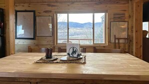 Ein Holzblockhaus-Interieur mit einem großen Esstisch, gerahmten Karten an den Wänden und Bergblick durch das Fenster.