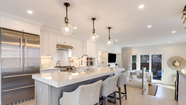 A modern kitchen with stainless steel appliances, a central island with bar stools, pendant lighting, and a view to a living area with a sofa and a TV.