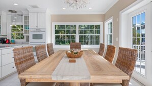 A spacious dining area with a large wooden table and wicker chairs, surrounded by large windows offering a view of lush greenery.