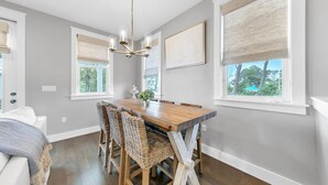 Un coin repas avec une table en bois et des chaises en osier, de grandes fenêtres équipées de stores et un lustre.
