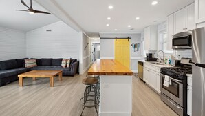 A modern kitchen with a central island, stainless steel appliances, and a dark-colored sofa in the living area. - 2 Incredible Units Equal Double the Amenities! Lazy Mermaid (Folly Beach)