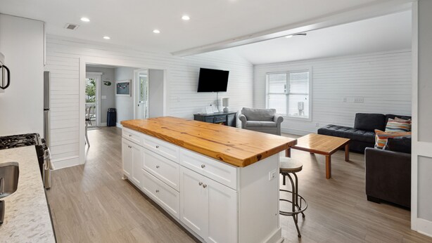 A modern kitchen with white cabinetry, a wooden countertop, and a built-in stove. There is a dining area with a table and chairs, and a living room with a sofa and a TV. - 2 Incredible Units Equal Double the Amenities! Lazy Mermaid (Folly Beach)