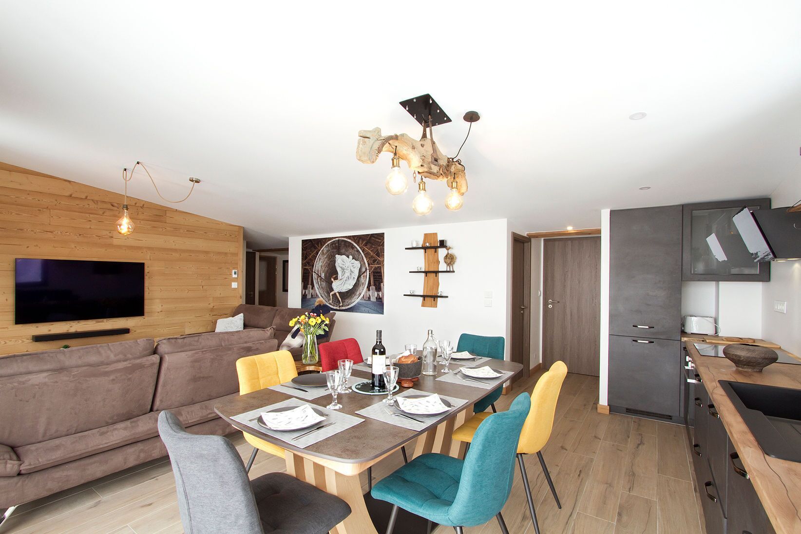 A modern living room with a flat-screen TV, wooden panel walls, a sofa, and a dining area with a wooden table and colorful chairs.