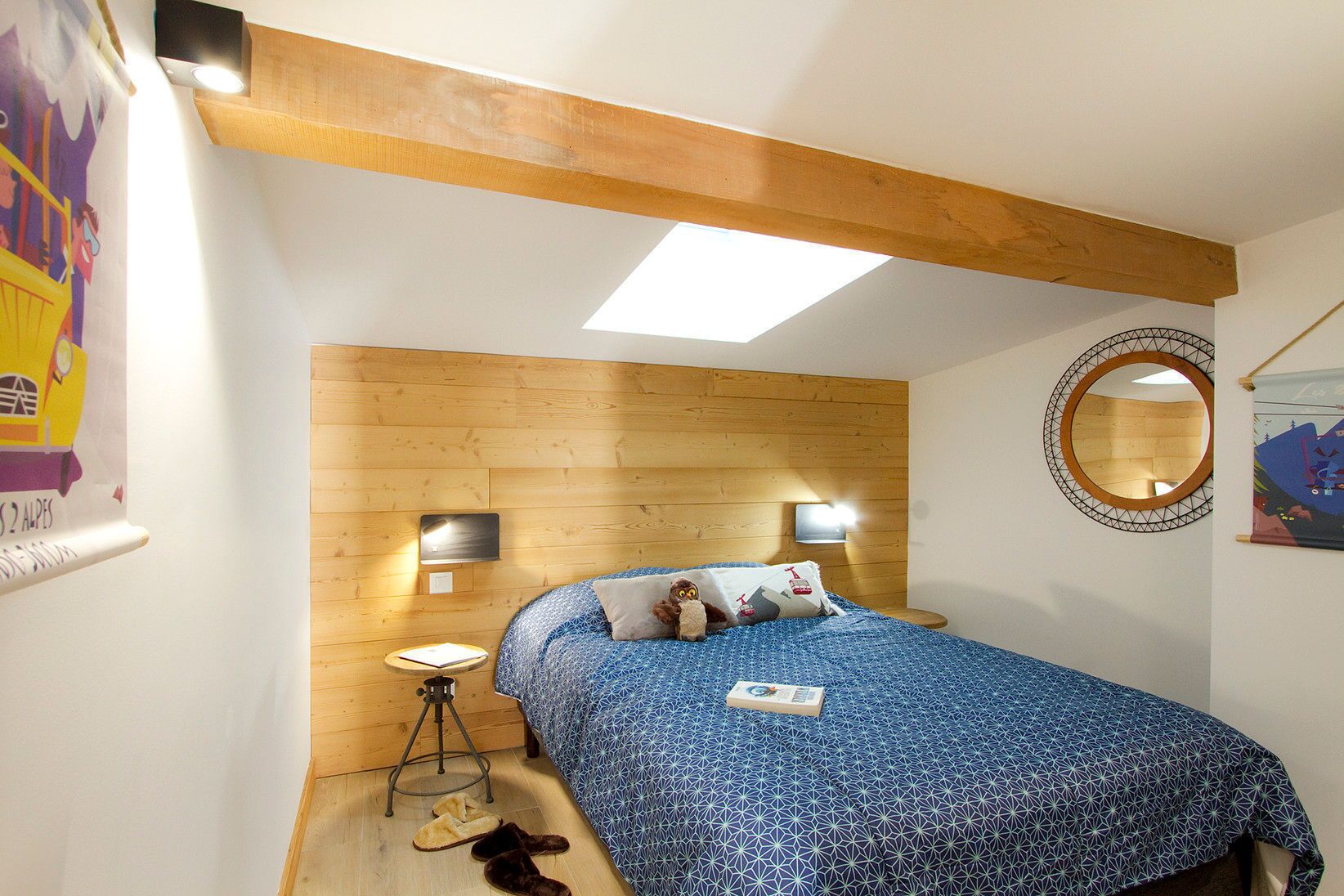 Un dormitorio con cabecera de madera, cama con colcha azul estampada, un espejo redondo y un taburete pequeño.