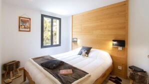 Un dormitorio con cabecera de madera, un ventanal grande, una cama con almohadas, una mesita de noche y zapatillas en el suelo.
