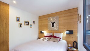 A bedroom with a wooden headboard, a bed with white bedding, red and beige pillows, a mounted TV, and a small wooden nightstand with a lamp. - Près du Télémixte du Diable | T2 + terrasse & Wifi (Les Deux Alpes)