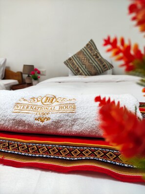 A neatly made bed with a white towel and a decorative pillow, a small bedside table with a lamp, and a vase with flowers. - Hostal International House Cusco (Cusco)