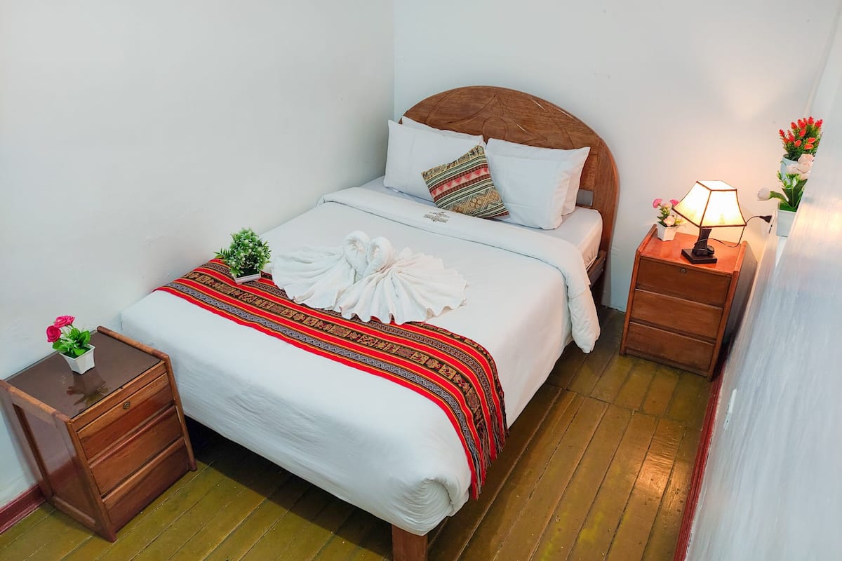 A neatly made bed with a white comforter and a red and black patterned blanket. There is a wooden nightstand with a lamp and a small potted plant beside the bed.