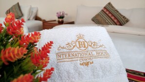 A hotel room with a white sofa, a decorative pillow, a small table with a vase of flowers, and a towel with the hotel name and logo. - Hostal International House Cusco (Cusco)