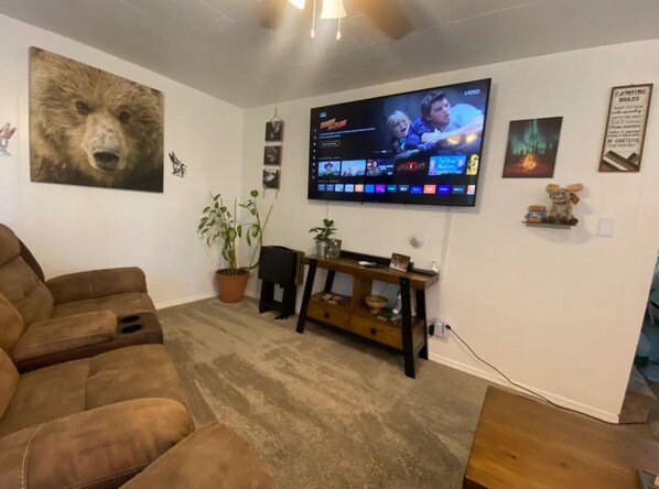 Una sala de estar con un televisor de pantalla plana grande montado en la pared, un sofá de cuero marrón, una mesita de madera y una planta en maceta.