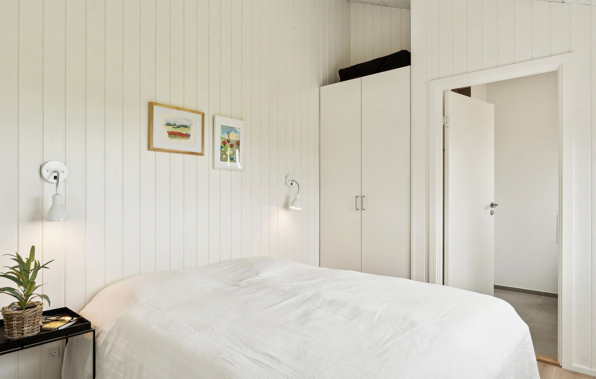 A bedroom with a bed, a nightstand with a plant, two wall-mounted lamps, and two framed pictures.