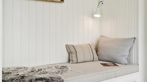 Eine weiße Wand mit Holzpaneelen, vor der eine weiße Bank steht. Eine weiße Lampe ist an der Wand befestigt und eine Felldecke liegt über der Bank.