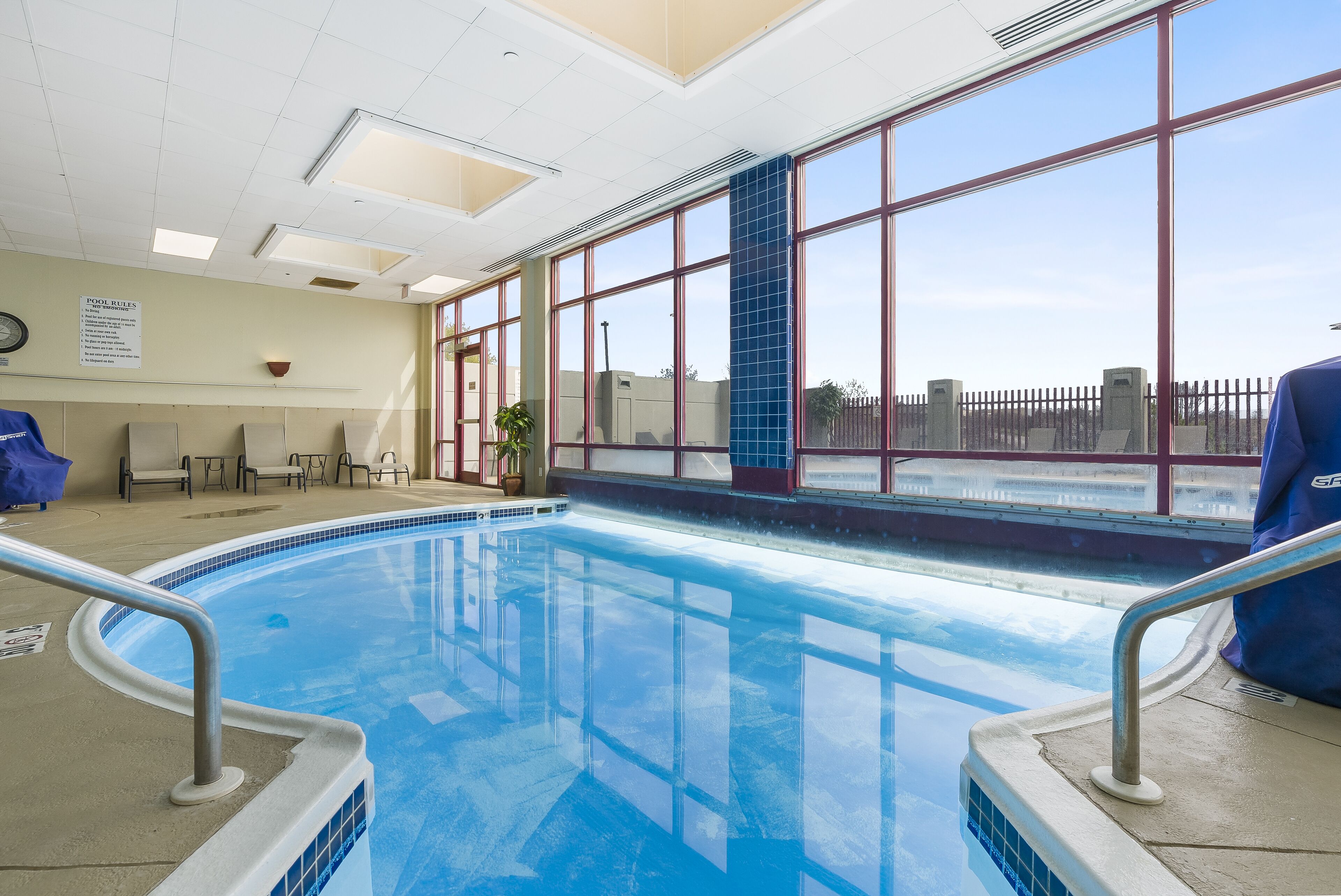An indoor swimming pool with clear blue water, a sunroof, and a view of a fence outside.