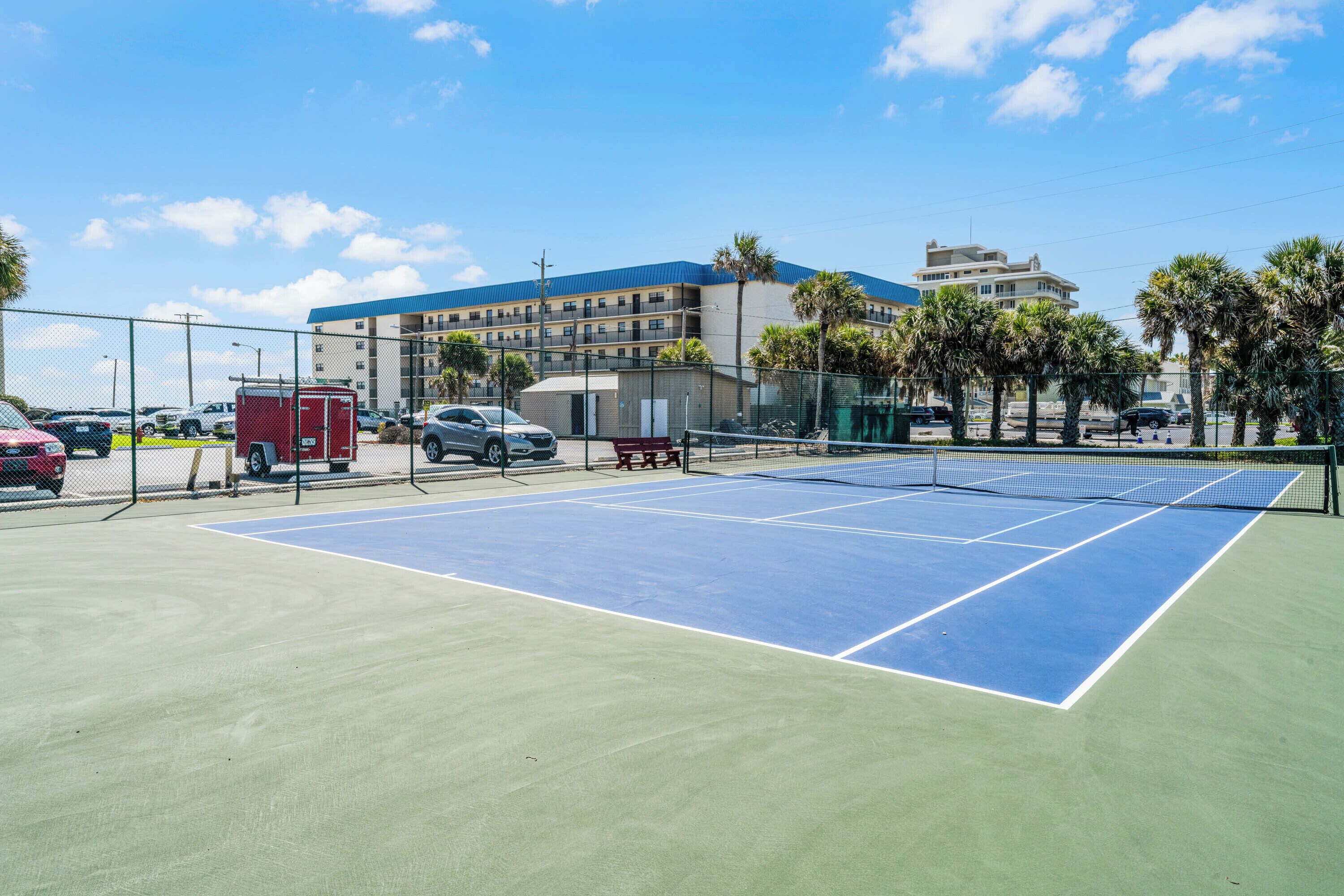 A tennis court with a net, surrounded by a fence, palm trees, and a building in the background.
