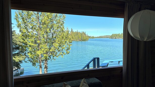 Ein Zimmer mit großem Fenster und Blick auf einen See, Holzeinrichtung sowie Bäume und einen Steg.