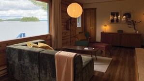 Ein gemütliches Zimmer mit großem Fenster und Seeblick, einer Holzbalkendecke, einem grünen Sofa, einem kleinen Tisch und einer Lampe.