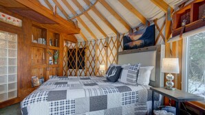 A cozy room with a large bed, wooden walls, and a window with a view of trees. - Unique 2Bds/1Bath Yurt | Kitchen, Wifi, Views! (Bozeman)
