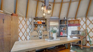 A rustic kitchen with a wooden table, shelves with dishes, a microwave, and a refrigerator. - Unique 2Bds/1Bath Yurt | Kitchen, Wifi, Views! (Bozeman)