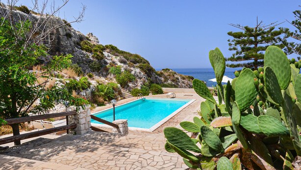 A pool area with a clear view of the sea, surrounded by greenery and rocky terrain.