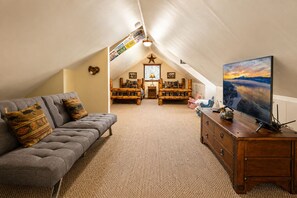 A cozy attic room with a gray sofa, wooden dresser, and a large TV. - Antelope Retreat - 5BR, Sauna, Foosball Table, Fireplace, Deck, BBQ (Big Bear City)