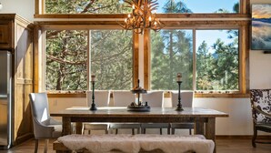 A dining area with a wooden table, chairs, and a bench. There is a chandelier above the table, and a large window with a view of trees. - Shem Shack - Modern, Game Room, Wet Bar, Fire Pit (Big Bear City)