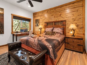 A bedroom with a wooden headboard, a bed with patterned bedding, a desk with a computer, a chair, a lamp, and a window with a view of trees. - Shem Shack | Luxury Mountain Retreat, Game Room, Wet Bar, Fire Pit & Views (Big Bear City)