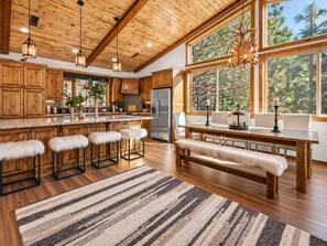 A spacious kitchen with wooden cabinets, a large island with bar stools, a built-in refrigerator, and a view of trees through large windows. - Shem Shack | Luxury Mountain Retreat, Game Room, Wet Bar, Fire Pit & Views (Big Bear City)