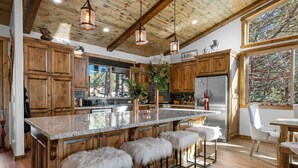 A spacious kitchen with wooden cabinets, a large island with bar stools, and a view of trees through the window. - Shem Shack - Modern, Game Room, Wet Bar, Fire Pit (Big Bear City)