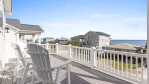 En træterrasse med hvide gelændere, hvide Adirondack-stole og udsigt til huse og havet.