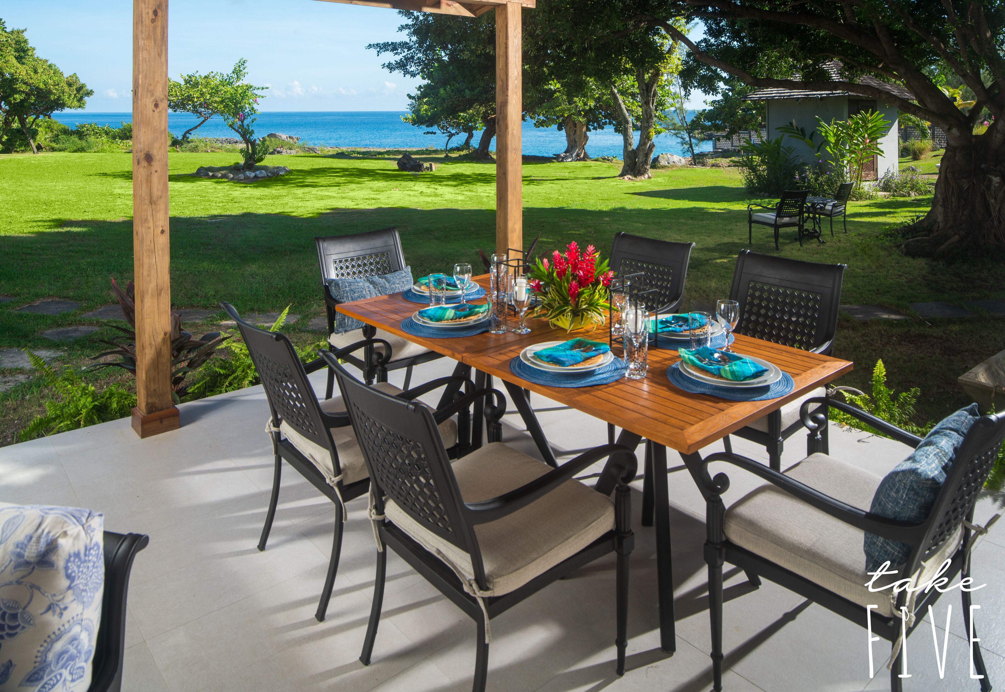Une table en bois, dressée pour un repas, entourée de chaises, avec vue sur l’océan et donnant sur un espace verdoyant parsemé d’arbres.