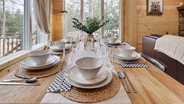 A dining table set with white plates, bowls, and cutlery, surrounded by woven placemats, with a vase of greenery in the center. - Mins to Skiing! Hot Tub-Fire Pit-Hammock-BBQ-Games (Lake Harmony)