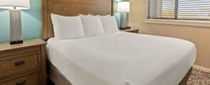 A hotel room with a large bed, a wooden headboard, a nightstand with a lamp, and a window with blinds.