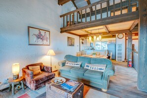 Una sala de estar acogedora con un sofá azul, un sillón marrón y una mesa de centro de mimbre. Se aprecia un comedor al fondo, con una cocina y una escalera que lleva a un nivel superior.