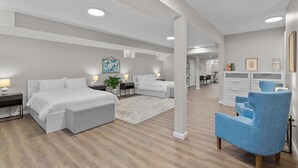 A modern hotel room with a large bed, a seating area with a blue armchair, and a kitchenette in the background.