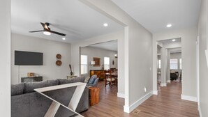 A spacious living room with a gray sectional sofa, wooden coffee table, and a ceiling fan. There is a dining area with a wooden table and chairs in the background.
