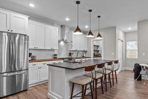 Uma cozinha moderna com armários brancos, eletrodomésticos de aço inoxidável, uma ilha central com banquetas, iluminação pendente e vista para a área de jantar adjacente.